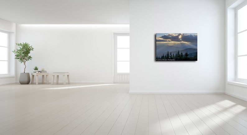 May include: A photograph of a mountain range with a sunbeam shining through the clouds. The mountain range is covered in trees and the sky is a light blue color.