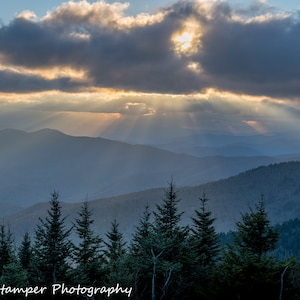 May include: A panoramic view of a mountain range with a cloudy sky. The sun is shining through the clouds, creating rays of light. The mountains are covered in trees and the sky is a light blue color.