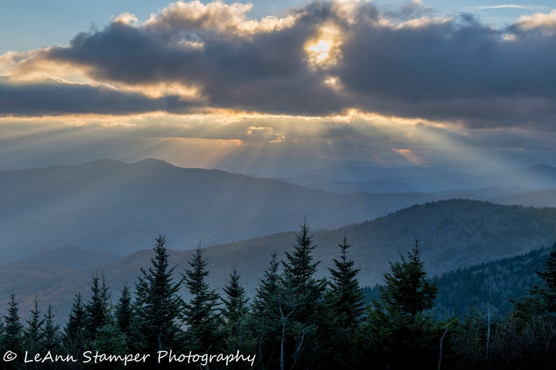 May include: A panoramic view of a mountain range with a cloudy sky. The sun is shining through the clouds, creating rays of light. The mountains are covered in trees and the sky is a light blue color.