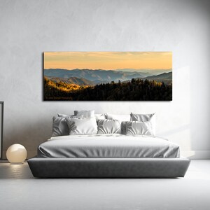 Puede incluir: Una fotograf&iacute;a panor&aacute;mica de una cordillera con una puesta de sol dorada en el fondo. Las monta&ntilde;as est&aacute;n cubiertas de &aacute;rboles y el cielo es de un azul suave. La imagen est&aacute; enmarcada y colgada en una pared sobre una cama en un dormitorio.