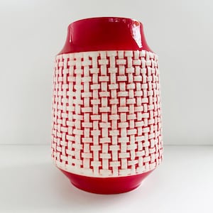 May include: A red and white ceramic vase with a woven pattern. The top and base are solid red, while the body features a textured white weave design. The vase has a cylindrical shape with a slightly flared top.