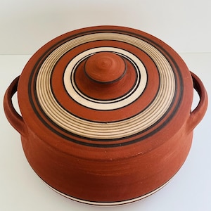 May include: A terracotta-colored ceramic cooking pot with a matching lid. The lid features concentric circles in black, white, and tan. The pot has two side handles for easy carrying. A classic kitchenware item.