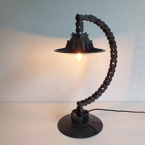 May include: A unique black desk lamp crafted from repurposed bicycle chain links. The lamp features a circular base, a curved chain arm, and a lampshade. The light bulb is visible, casting a warm glow. The lamp is plugged into a wall outlet.