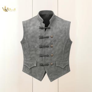 Op de afbeelding: Een grijze mouwloze vest met een opstaande kraag en decoratieve knevelsluitingen. De vest heeft twee voorzakken en bronskleurige knopen. Het logo "WEAR SHOP" staat in de linkerbovenhoek.