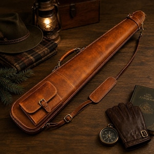 May include: A long, tan leather carrying case with a structured design, featuring a top handle, an adjustable shoulder strap with a padded section, and a small exterior pocket secured with a buckle strap and metal hardware on the front.
