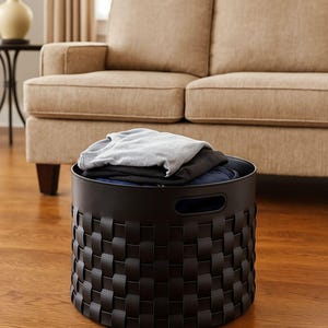 May include: A black woven storage basket filled with folded clothes. The round basket has a handle on each side and sits on a hardwood floor. A beige sofa is in the background.