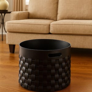 May include: A black woven basket with a cylindrical shape, featuring a repeating pattern of interwoven strips. The basket has two handle cutouts and sits on a hardwood floor. A beige sofa is in the background.
