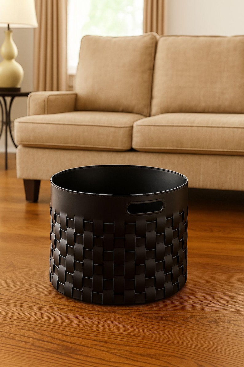 May include: A black woven basket with a cylindrical shape, featuring a repeating pattern of interwoven strips. The basket has two handle cutouts and sits on a hardwood floor. A beige sofa is in the background.