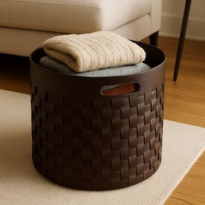 Puede incluir: Una cesta de almacenamiento tejida de color marrón oscuro con forma redonda y dos asas. La cesta contiene una manta beige doblada y una manta gris. La cesta está sobre una alfombra de color claro.