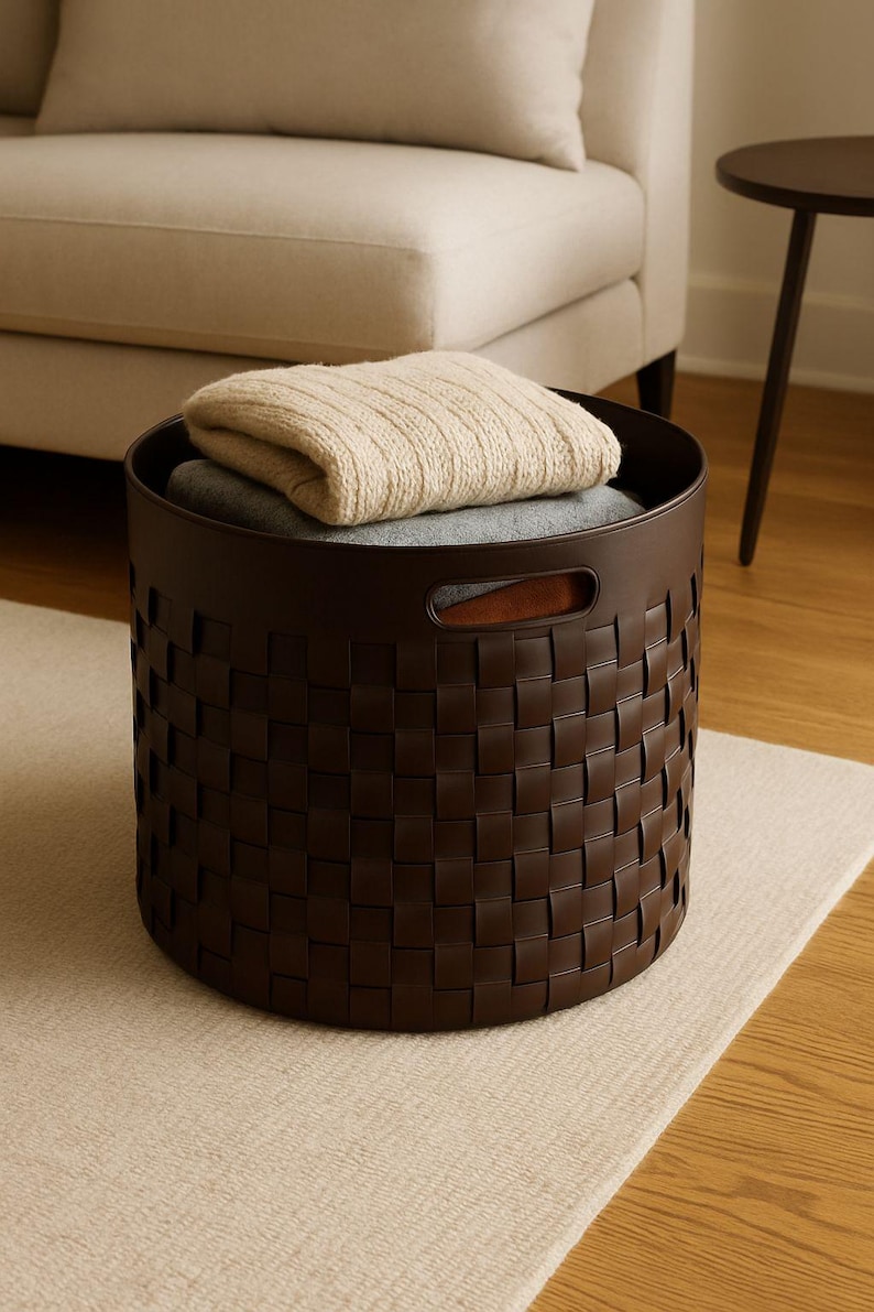 May include: A dark brown woven storage basket with a round shape and two handles. The basket contains a folded beige blanket and a gray blanket. The basket is sitting on a light-colored rug.