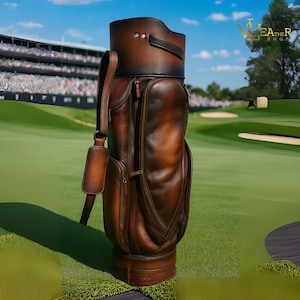 May include: A brown leather golf bag with a shoulder strap and multiple pockets. The bag has a dark brown top and base, with a lighter brown body. The bag is on a green golf course.
