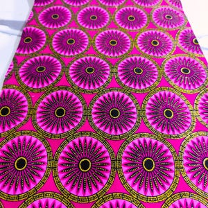 Könnte beinhalten: Leuchtend pinkfarbener Stoff mit einem sich wiederholenden Muster aus großen, kreisförmigen Blumenmustern. Jedes Motiv hat eine schwarze Mitte, strahlende rosa Blütenblätter und einen gold-schwarzen Rand. Der Stoff ist gefaltet und zeigt seine Textur.