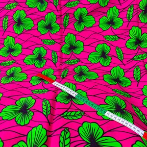 May include: Bright pink fabric with a repeating pattern of green shamrock-like leaves with black outlines. A white and green measuring tape is laid across the fabric, showing measurements in inches.