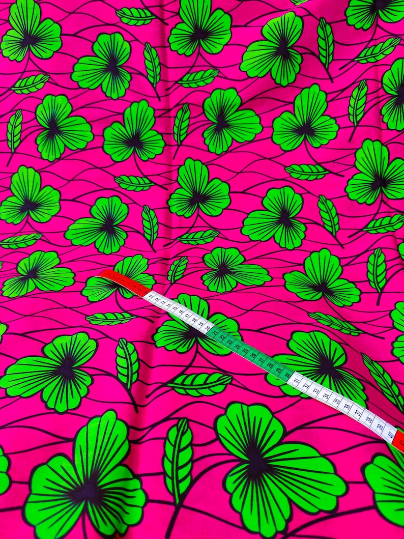 May include: Bright pink fabric with a repeating pattern of green shamrock-like leaves with black outlines. A white and green measuring tape is laid across the fabric, showing measurements in inches.