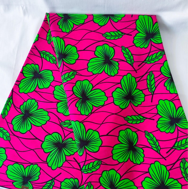 May include: Bright pink fabric with a repeating pattern of green shamrock-like flowers and leaves. The flowers have black centers and are outlined in green. The background features thin, wavy black lines.