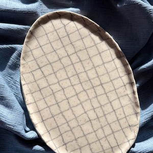 Pode incluir: Um prato oval de cerâmica esbranquiçada com um padrão de grade em cinza escuro. O prato está sobre um fundo de tecido azul, criando um contraste. Provavelmente usado para servir alimentos.