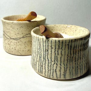 May include: Two cylindrical ceramic bowls with speckled cream glaze and wooden spoons. The front bowl features vertical black line patterns, while the back bowl displays a horizontal wavy black line design. Both bowls have a wide, open top.