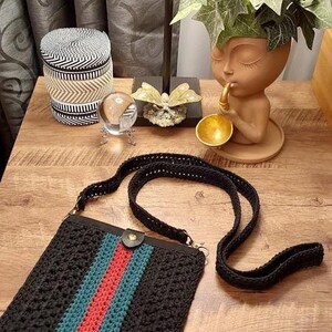 May include: Black crochet crossbody bag with a red and teal stripe design. The bag has a black strap and a leather button closure. The bag is sitting on a wooden surface.