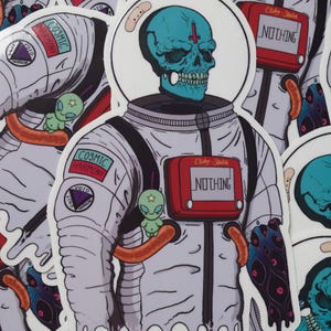 May include: A collection of stickers featuring an astronaut with a teal skull head and a red device that says "NOTHING." The astronaut's suit has patches that say "COSMIC HARMONY" and a small green alien is attached to the suit.