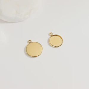 Puede incluir: Dos colgantes redondos de color dorado con peque&ntilde;os lazos para sujetarlos a una cadena o cord&oacute;n. Un colgante tiene una superficie plana, mientras que el otro tiene un &aacute;rea empotrada para agregar un dise&ntilde;o o imagen. Los colgantes se muestran sobre una superficie blanca.
