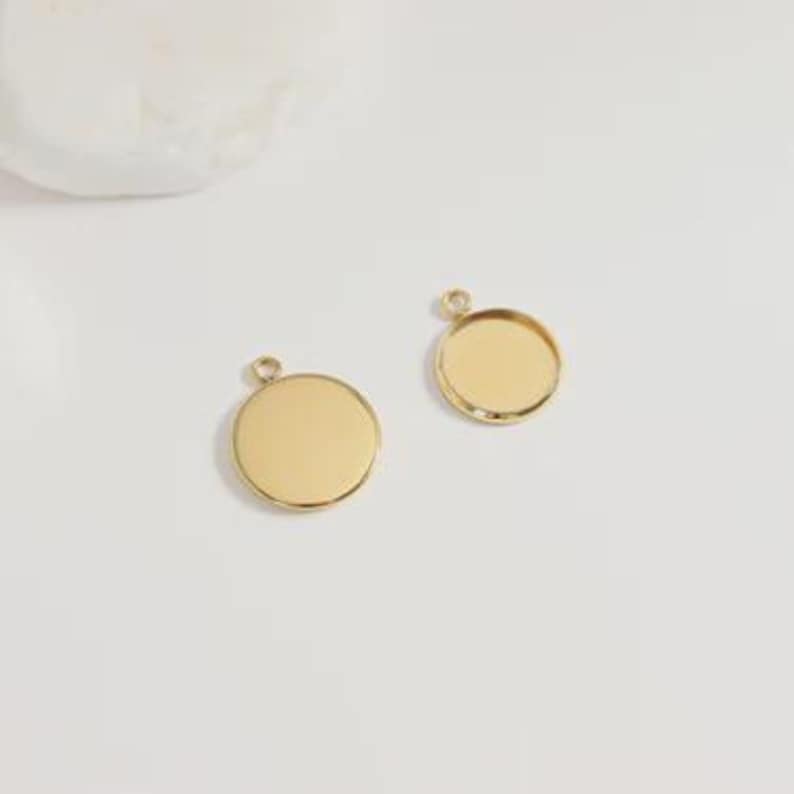 Puede incluir: Dos colgantes redondos de color dorado con peque&ntilde;os lazos para sujetarlos a una cadena o cord&oacute;n. Un colgante tiene una superficie plana, mientras que el otro tiene un &aacute;rea empotrada para agregar un dise&ntilde;o o imagen. Los colgantes se muestran sobre una superficie blanca.