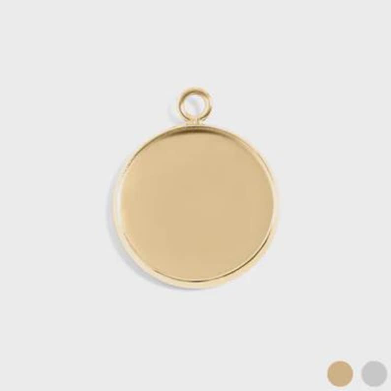 Puede incluir: Un colgante redondo de color dorado con una superficie lisa y plana y un peque&ntilde;o lazo en la parte superior para sujetarlo a una cadena. El colgante se muestra sobre un fondo neutro, con una paleta de colores dorados, beige y grises.