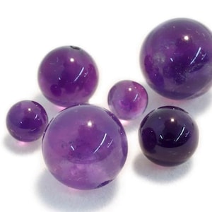 May include: A collection of six amethyst spheres of varying sizes, ranging from small to large, with a purple hue.