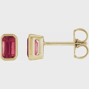 May include: A pair of gold stud earrings featuring rectangular, faceted, red gemstones. The gemstones are set within a gold bezel. The earrings have a post and butterfly backing. The earrings are displayed against a neutral background.