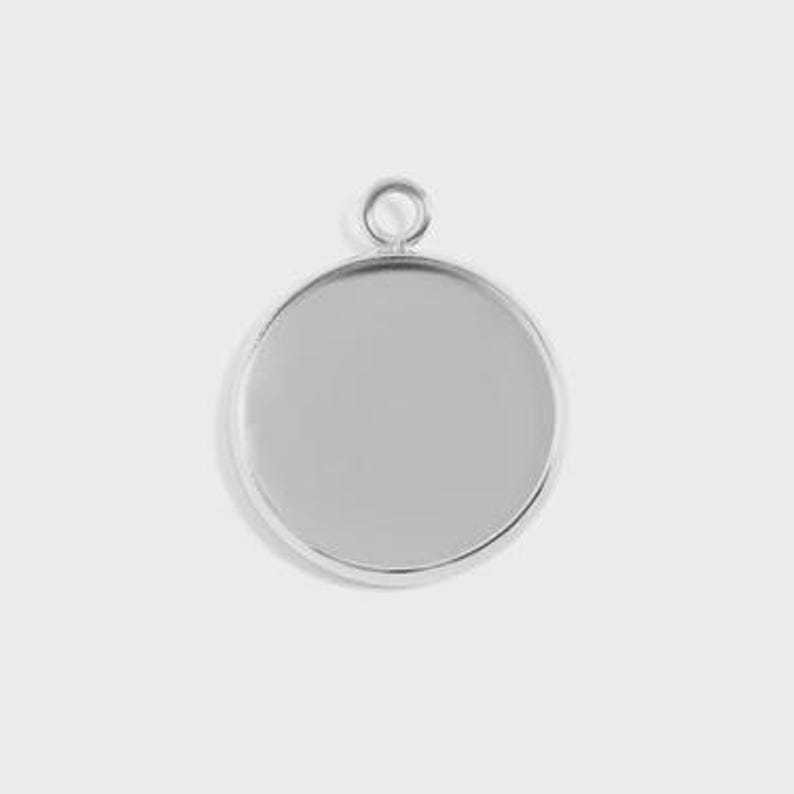 Puede incluir: Un colgante redondo de color plateado con un bisel elevado y un peque&ntilde;o lazo en la parte superior para sujetarlo a una cadena o cord&oacute;n. El colgante tiene una superficie lisa y reflectante, perfecto para a&ntilde;adir un dise&ntilde;o o imagen personalizado. El colgante se muestra sobre un fondo liso y claro.