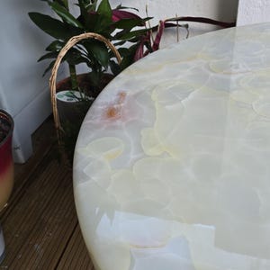 May include: A close-up of a round, off-white table with a glossy finish. The table's surface has a marbled pattern with light yellow and cream tones. The table is near potted plants and a wooden deck.