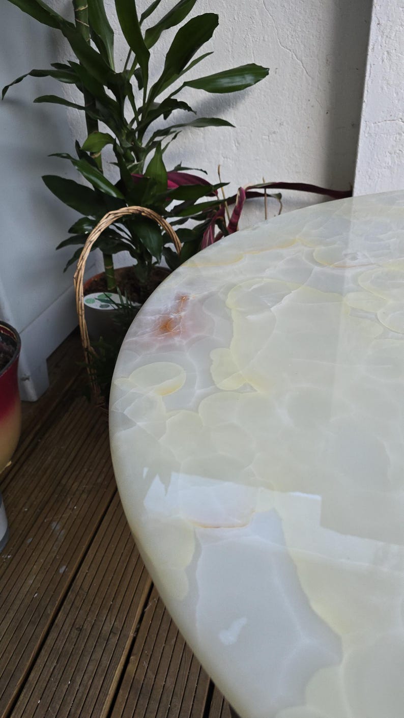May include: A close-up of a round, off-white table with a glossy finish. The table's surface has a marbled pattern with light yellow and cream tones. The table is near potted plants and a wooden deck.