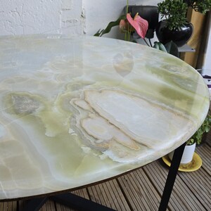 May include: A round coffee table with a polished, light green onyx top. The surface has a marbled pattern with white and tan veining. The table has a black metal frame and sits on a wooden deck.