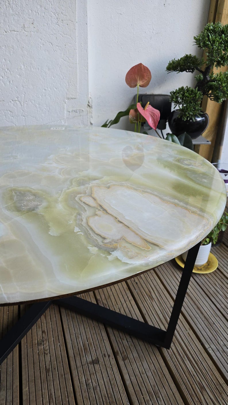 May include: A round coffee table with a polished, light green onyx top. The surface has a marbled pattern with white and tan veining. The table has a black metal frame and sits on a wooden deck.