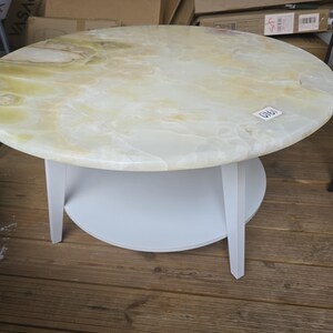 May include: A round coffee table with a light-colored, marbled top and a white base. The table has a circular shelf and three legs. The top appears to be made of onyx or a similar stone. The table is on a wooden floor.