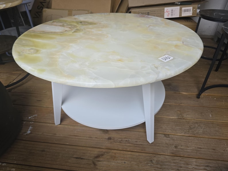 May include: A round coffee table with a light-colored, marbled top and a white base. The table has a circular shelf and three legs. The top appears to be made of onyx or a similar stone. The table is on a wooden floor.