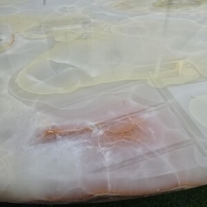 May include: A close-up of a round table with a polished, translucent surface. The table's surface features a swirling pattern of white, cream, and light brown hues, resembling natural stone or marble. The table is set on a green surface.