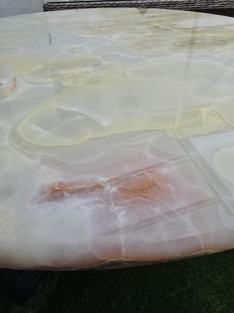 May include: A close-up of a round table with a polished, translucent surface. The table's surface features a swirling pattern of white, cream, and light brown hues, resembling natural stone or marble. The table is set on a green surface.