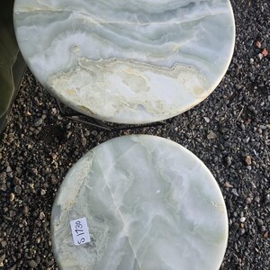 May include: Two round, light green and white marble table tops with unique veining patterns. The tops have a polished finish and are displayed outdoors on a gravel surface. Each top has a price tag attached.