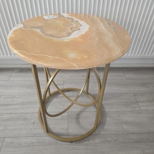 Op de afbeelding: Een ronde bijzettafel met een marmeren blad in beige en witte tinten. De tafel heeft een goudkleurig metalen onderstel met een ingewikkeld ontwerp. De tafel is ongeveer 50 cm in diameter en 61 cm hoog.