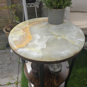 May include: A small, round side table with a white and yellow onyx top. The table has two wooden shelves and a dark metal frame. A small potted plant sits on the table's surface.