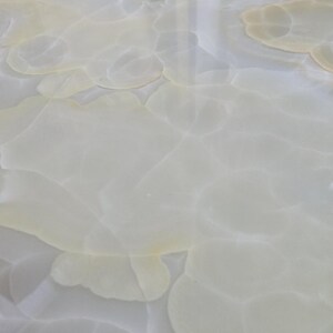 May include: Close-up of a polished, light-colored stone surface with a swirling, cloud-like pattern. The stone has a translucent quality, with subtle variations in color, ranging from white to pale yellow. The overall effect is smooth and elegant.