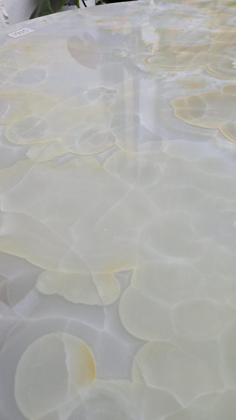 May include: Close-up of a polished, light-colored stone surface with a swirling, cloud-like pattern. The stone has a translucent quality, with subtle variations in color, ranging from white to pale yellow. The overall effect is smooth and elegant.