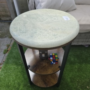 Puede incluir: Una mesa auxiliar redonda con una tapa texturizada de color verde claro y dos estantes inferiores. La mesa tiene una estructura de metal oscuro y un Cubo de Rubik está en el estante inferior. La mesa está sobre una superficie verde.
