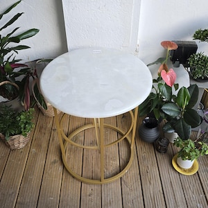 Può includere: Un tavolino rotondo con piano in marmo bianco e base in metallo dorato. Il tavolo è circondato da piante in vaso, creando un ambiente naturale e decorativo. Il design del tavolo è semplice ma elegante. Il diametro è di circa 40 cm.