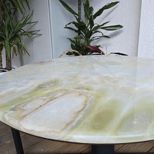 May include: A round table with a polished, light green and white onyx top. The table has a black metal base and is set against a backdrop of plants and a white wall.