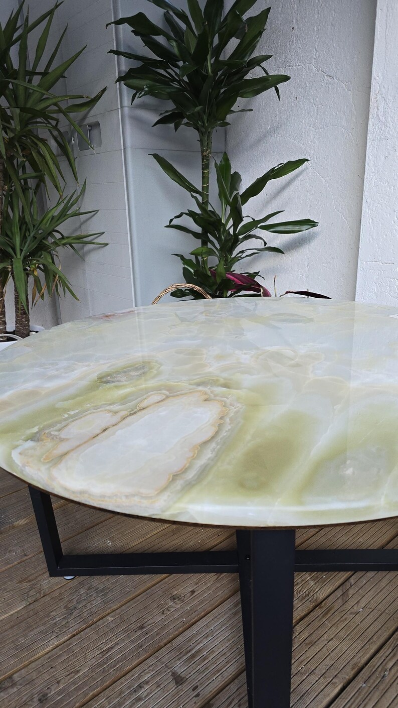May include: A round table with a polished, light green and white onyx top. The table has a black metal base and is set against a backdrop of plants and a white wall.