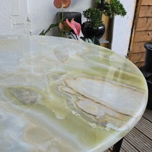 May include: Oval-shaped onyx table with a polished surface. The table features a mix of white, green, and tan colors, with a black metal base. The table is a decorative piece for a living room or patio.
