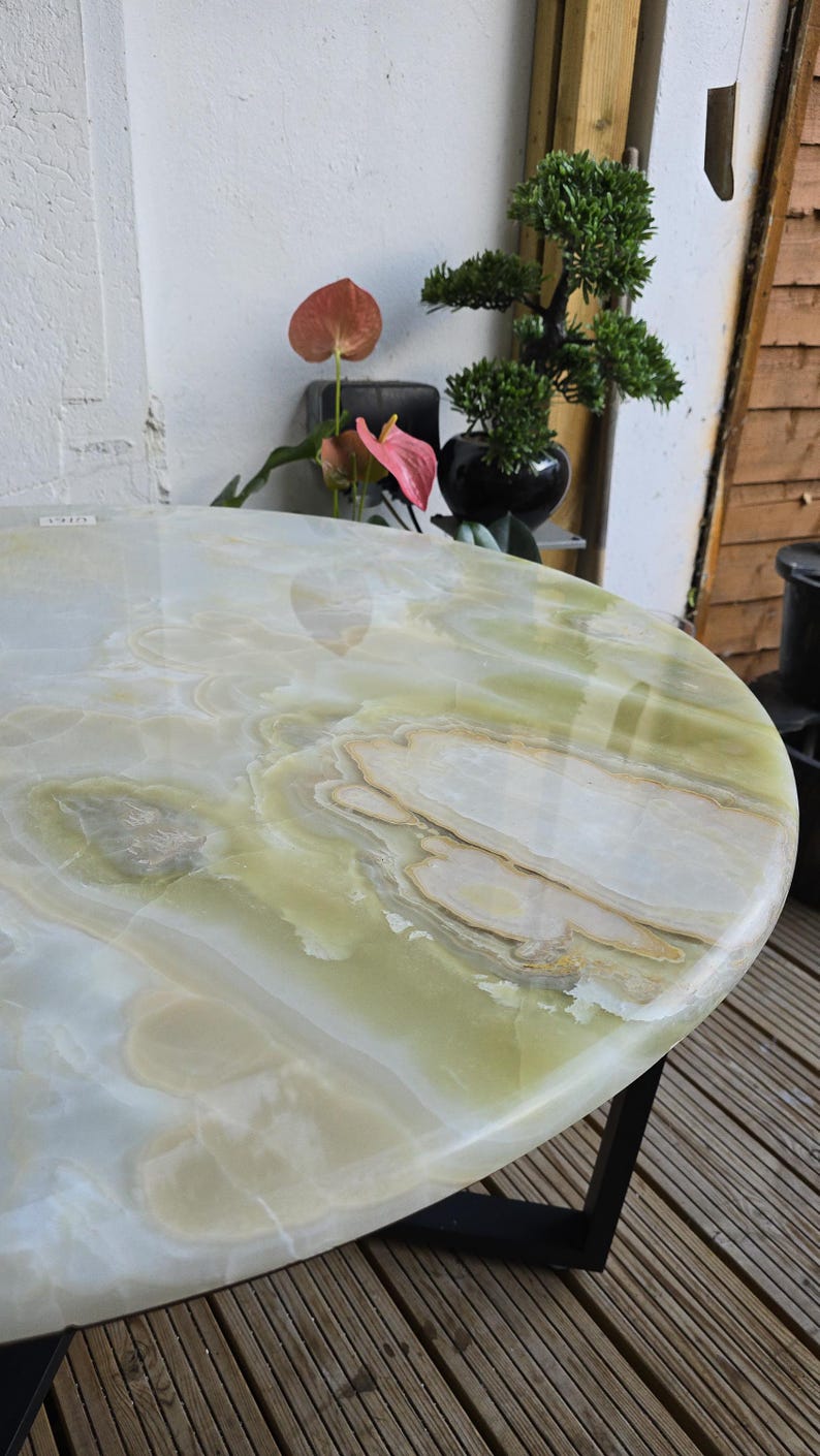 May include: Oval-shaped onyx table with a polished surface. The table features a mix of white, green, and tan colors, with a black metal base. The table is a decorative piece for a living room or patio.