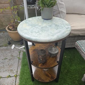May include: A three-tiered side table with a round, light green and white patterned top. The table has two lower shelves made of wood and a black metal frame. A small potted plant sits on top.