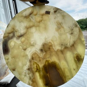 May include: A large, round, translucent stone slab with a unique pattern of light yellow, brown, and white swirls. The stone has a polished surface and a natural, organic appearance. The number "1910" is visible on a small sticker.